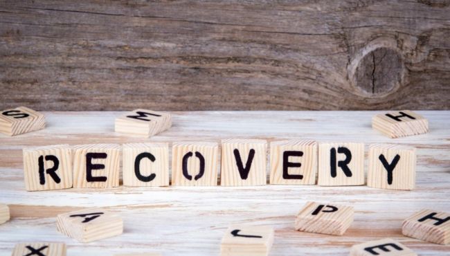 the word recovery spelled out with scrabble tiles in front a piece of wood